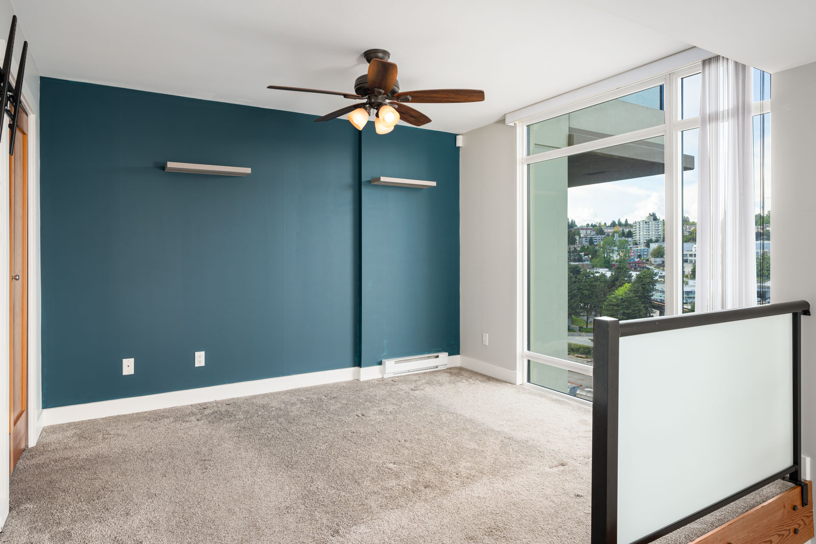 Empty room with blue accent wall, ceiling fan, and large window letting in natural light, overlooking a cityscape. Carpeted floor and modern railing are visible.