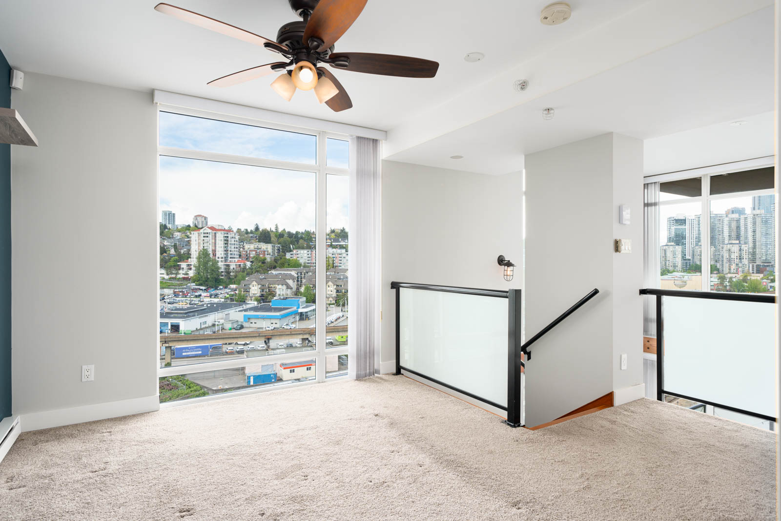Bright corner room with beige carpet, ceiling fan, large window overlooking city buildings, and staircase with glass railing.