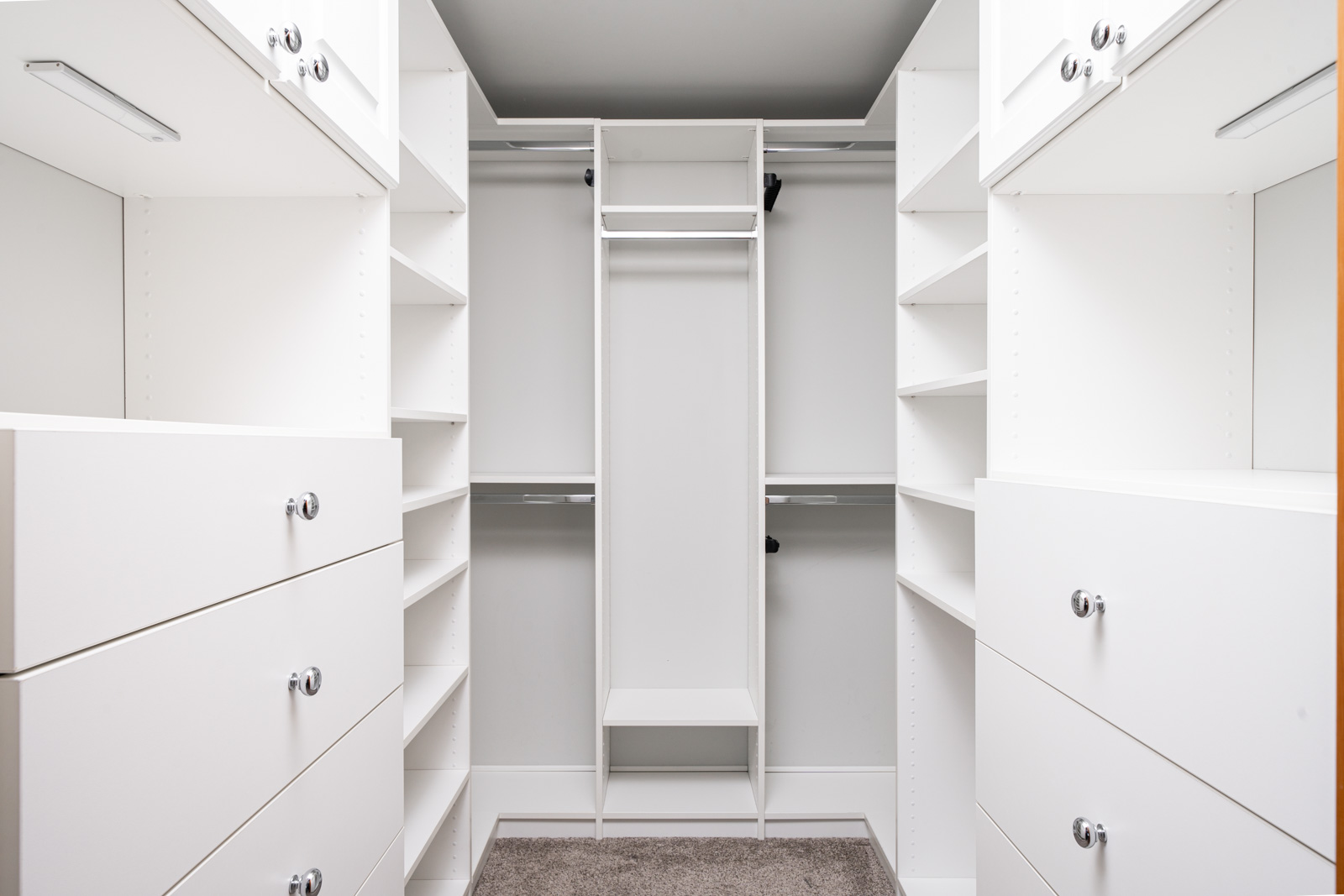 Walk-in closet with white built-in shelves, drawers, and hanging rods, all empty, with beige carpet flooring and overhead lighting.