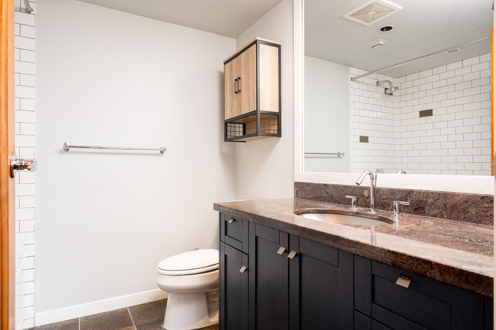 Modern bathroom with dark cabinets, a granite countertop, wall-mounted cabinet, toilet, towel bar, and a walk-in shower with subway tile visible in the mirror.