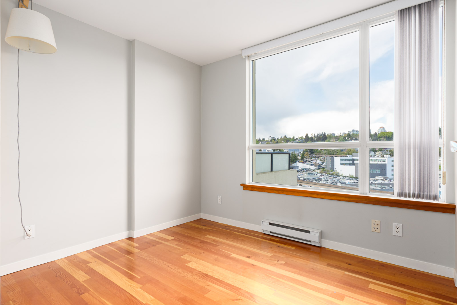 Unfurnished room with light gray walls, wood flooring, a large window with city view, and a lamp on the left wall.