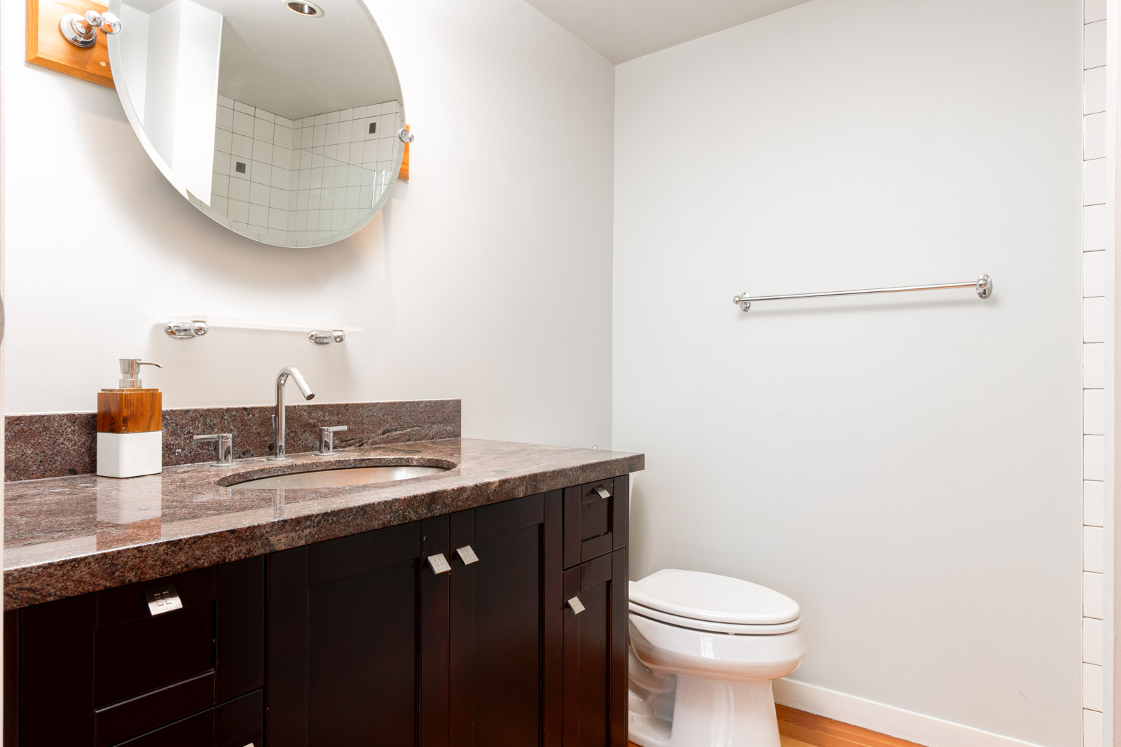 Modern bathroom with a dark wood vanity, granite countertop, round mirror, white walls, toilet, and a towel bar.