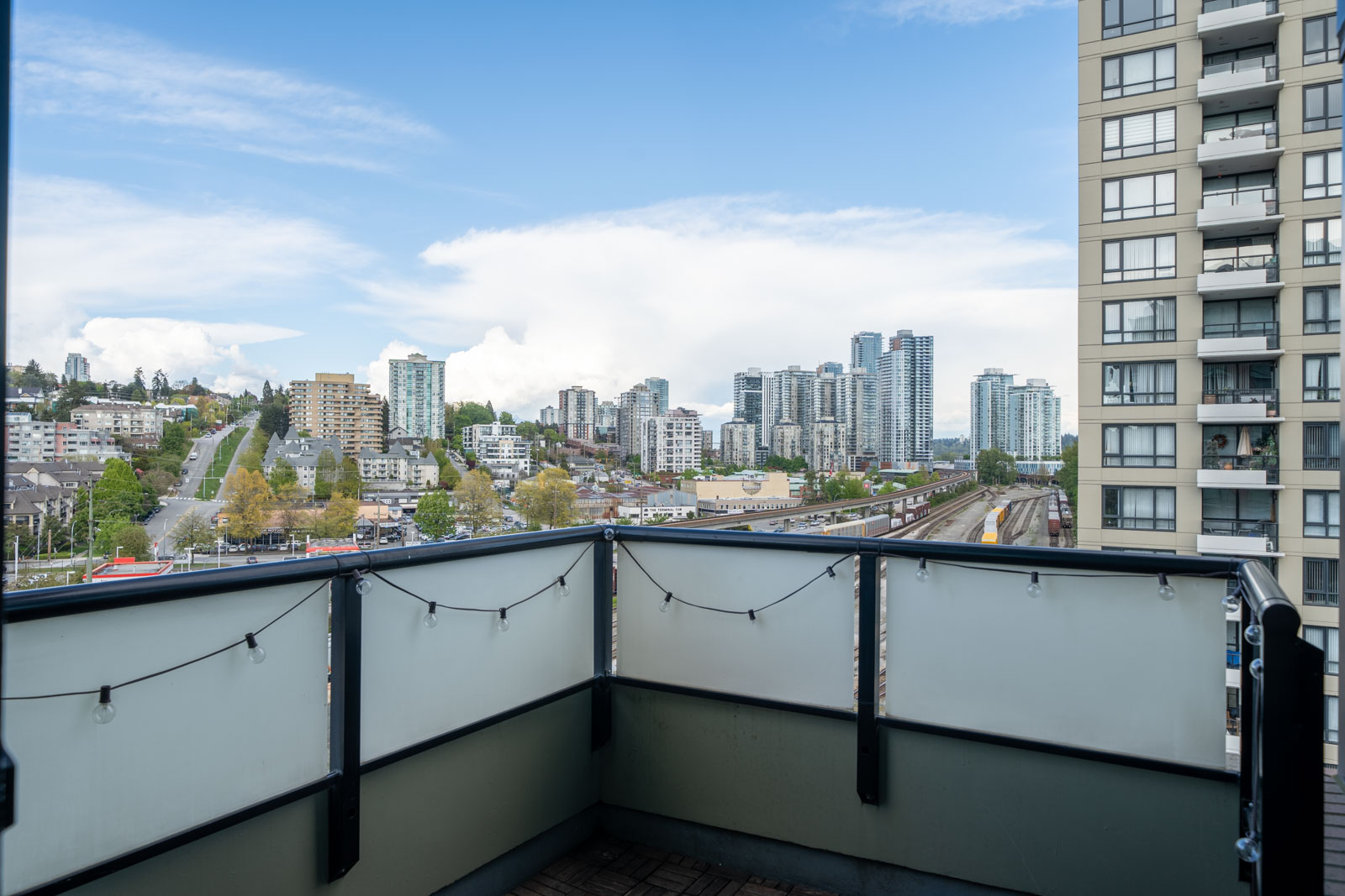 A cityscape view from a balcony with glass railings, showing high-rise buildings, a roadway, and a partly cloudy sky.