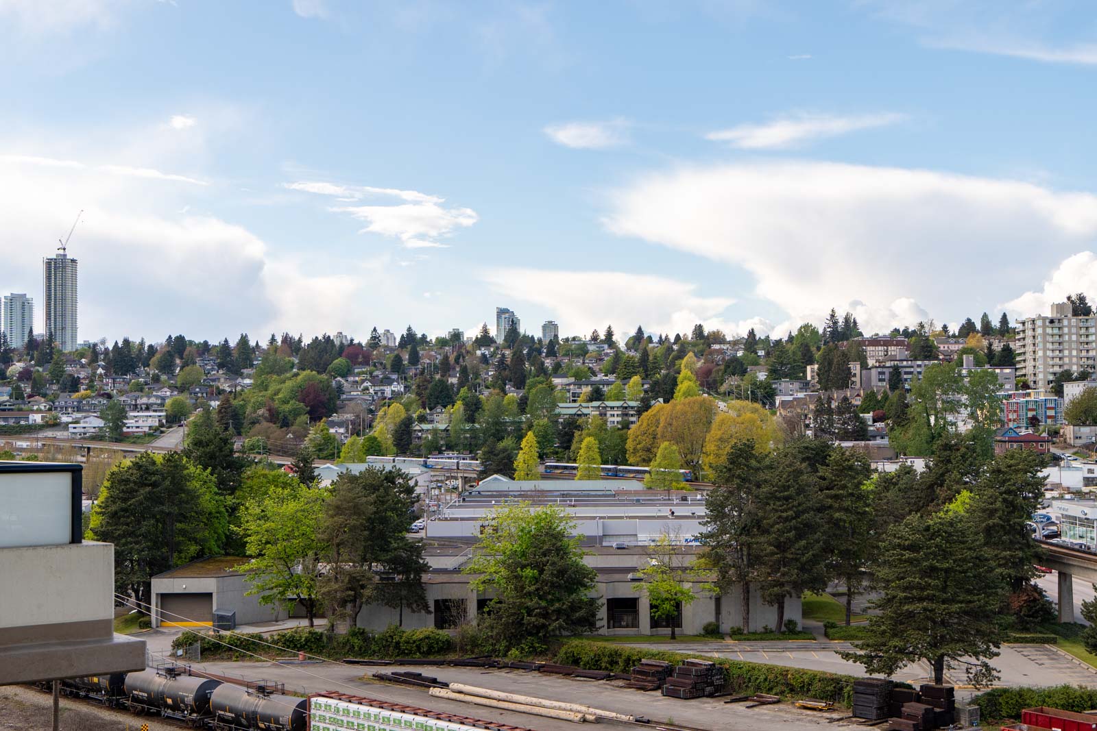 Urban landscape with industrial buildings in the foreground, residential houses and trees on a hillside in the background, and a few high-rise buildings under a partly cloudy sky.