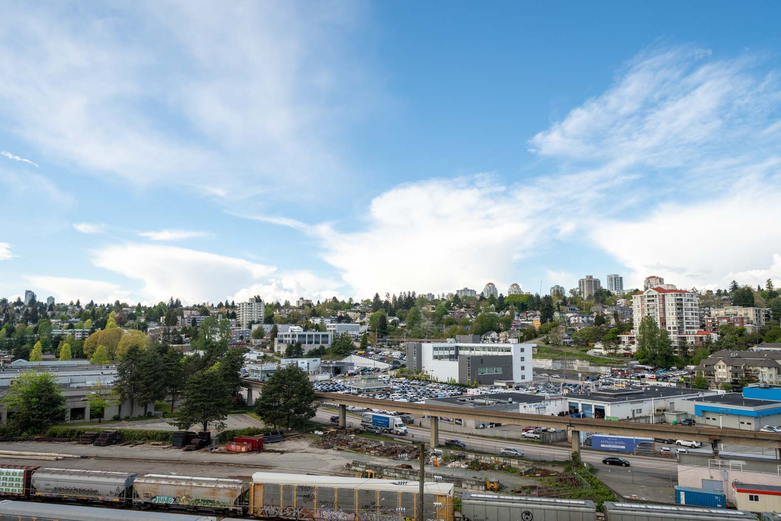 Urban landscape with commercial buildings, a parking lot, construction area, train tracks, and residential buildings under a partly cloudy sky.