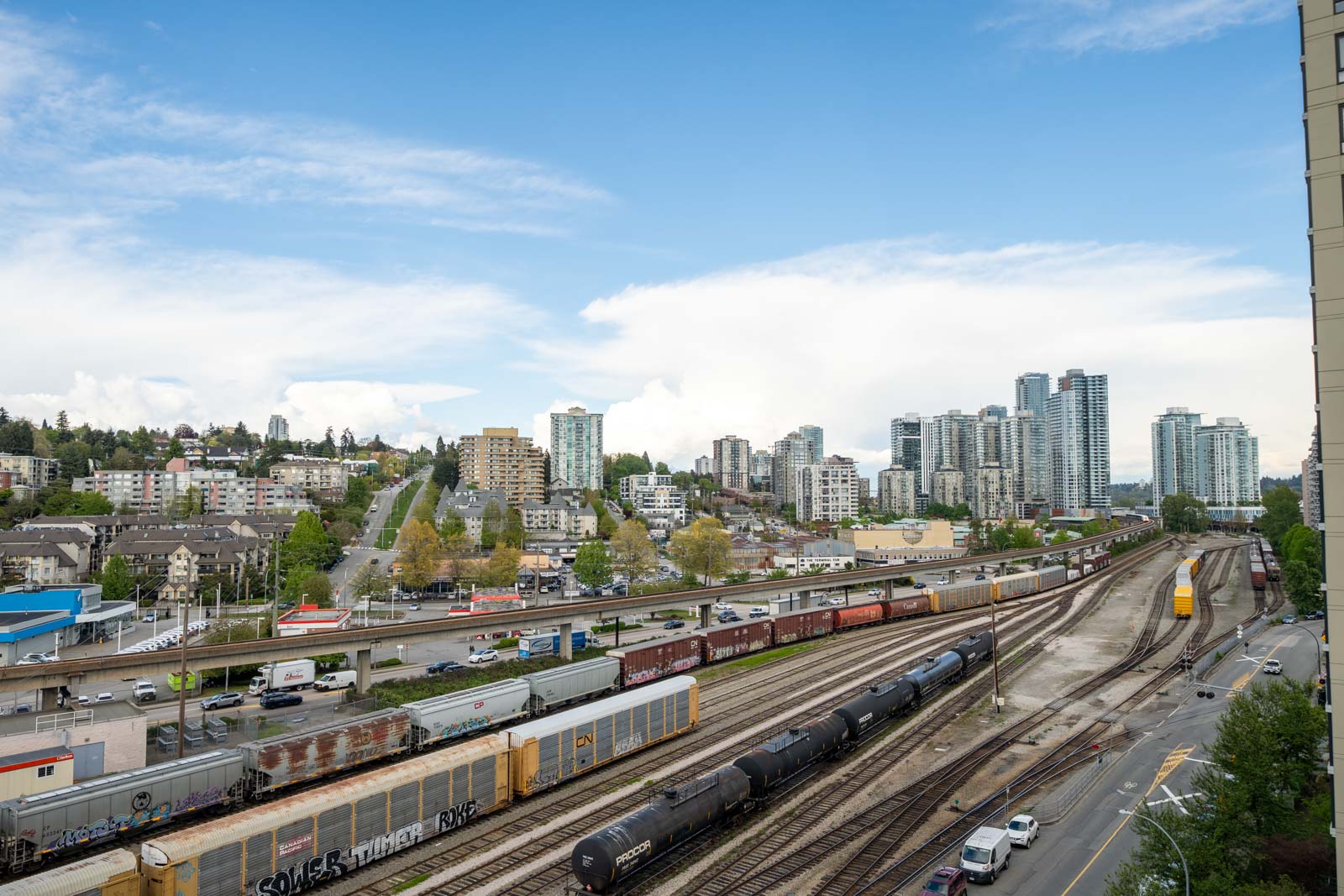 A cityscape with multiple train tracks, parked freight trains, roads with cars, and high-rise buildings under a partly cloudy sky.
