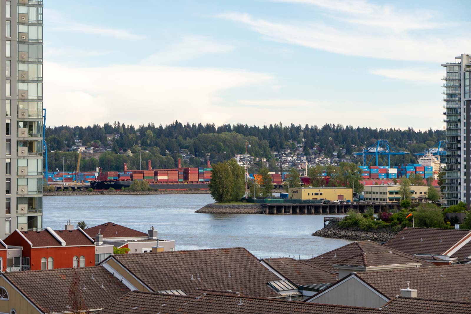 View of a river with shipping containers on the opposite bank, framed by apartment buildings and rooftops in the foreground, and trees in the background.