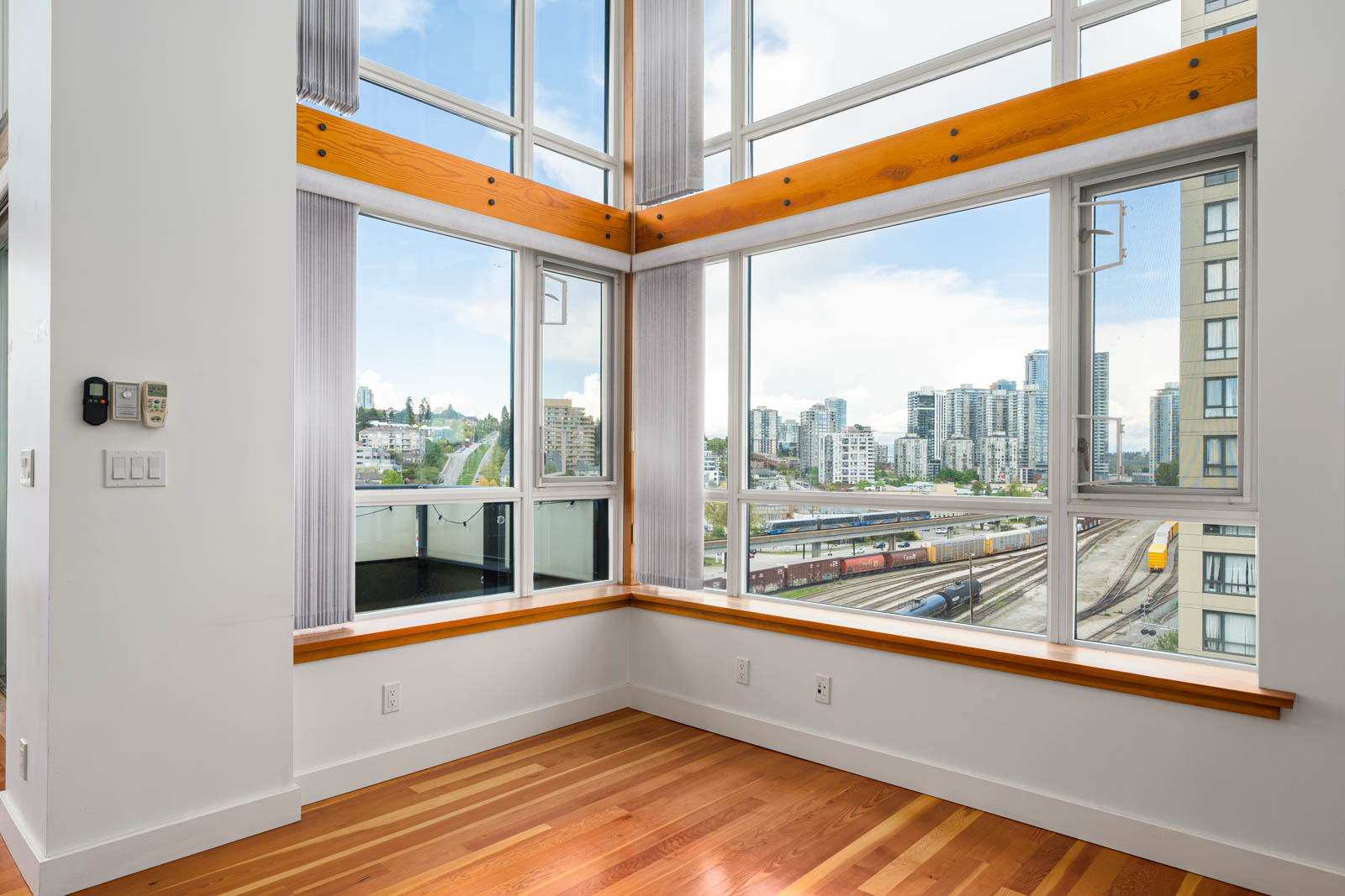 Corner of a modern room with large floor-to-ceiling windows, wood floors, white walls, and a cityscape view featuring buildings, roads, and train tracks.