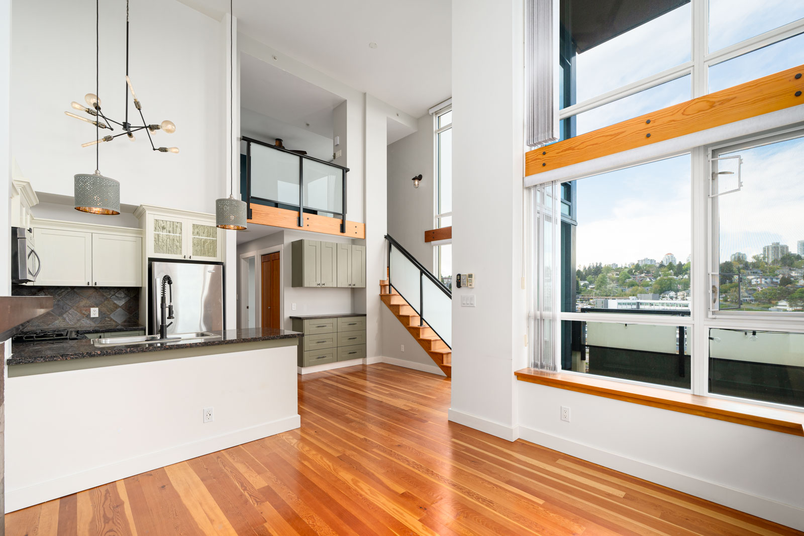 Modern open-plan loft with hardwood floors, large windows, a kitchen with pendant lights, and a staircase leading to an upper level.