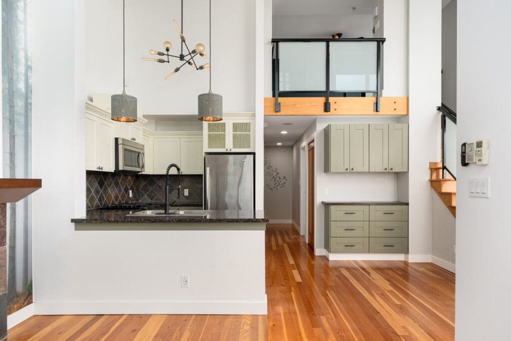 Modern kitchen with white cabinets, stainless steel appliances, granite countertops, pendant lights, hardwood floors, and a hallway leading to stairs with glass railing.