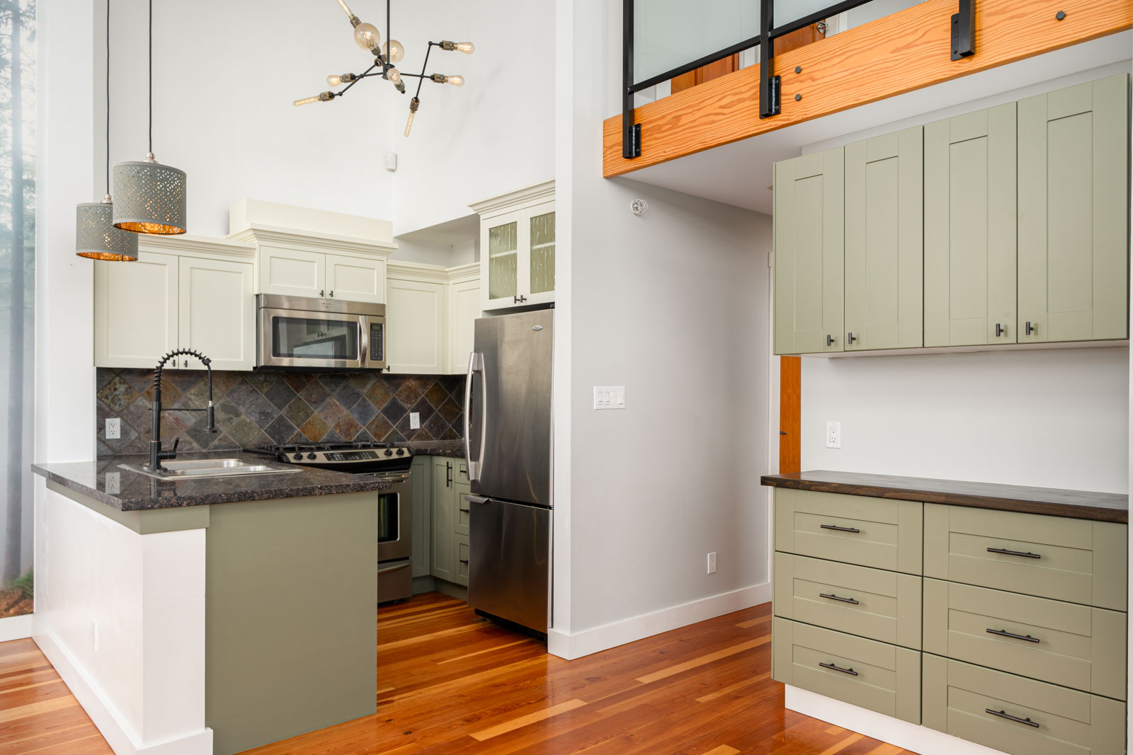 Modern kitchen with light green cabinets, stainless steel appliances, dark countertops, pendant lighting, and wood flooring. Upper loft with railing visible above cabinetry.