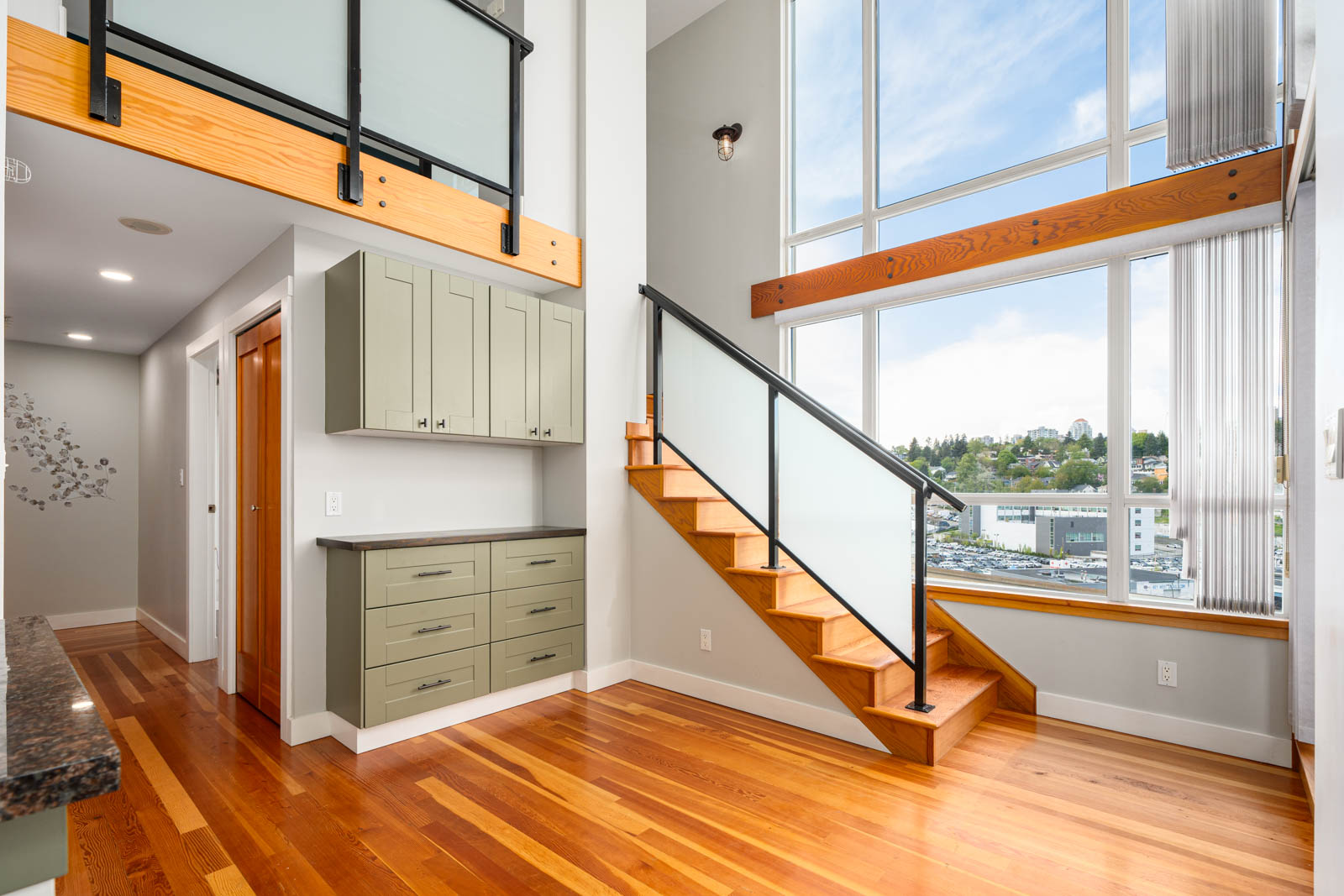Modern interior with large windows, wooden floors, light green cabinets, and a staircase with glass railing leading to an upper loft area.