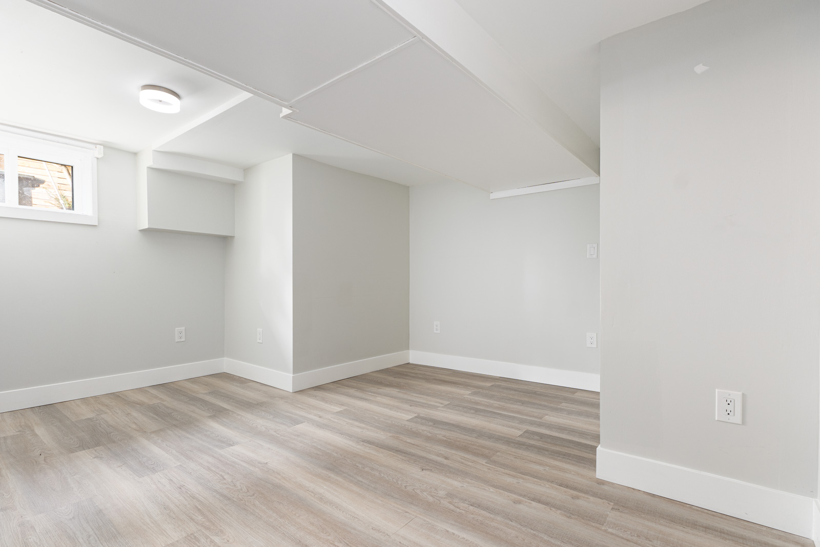 A clean, empty basement room with light gray walls, laminate wood flooring, a small window, and multiple electrical outlets.