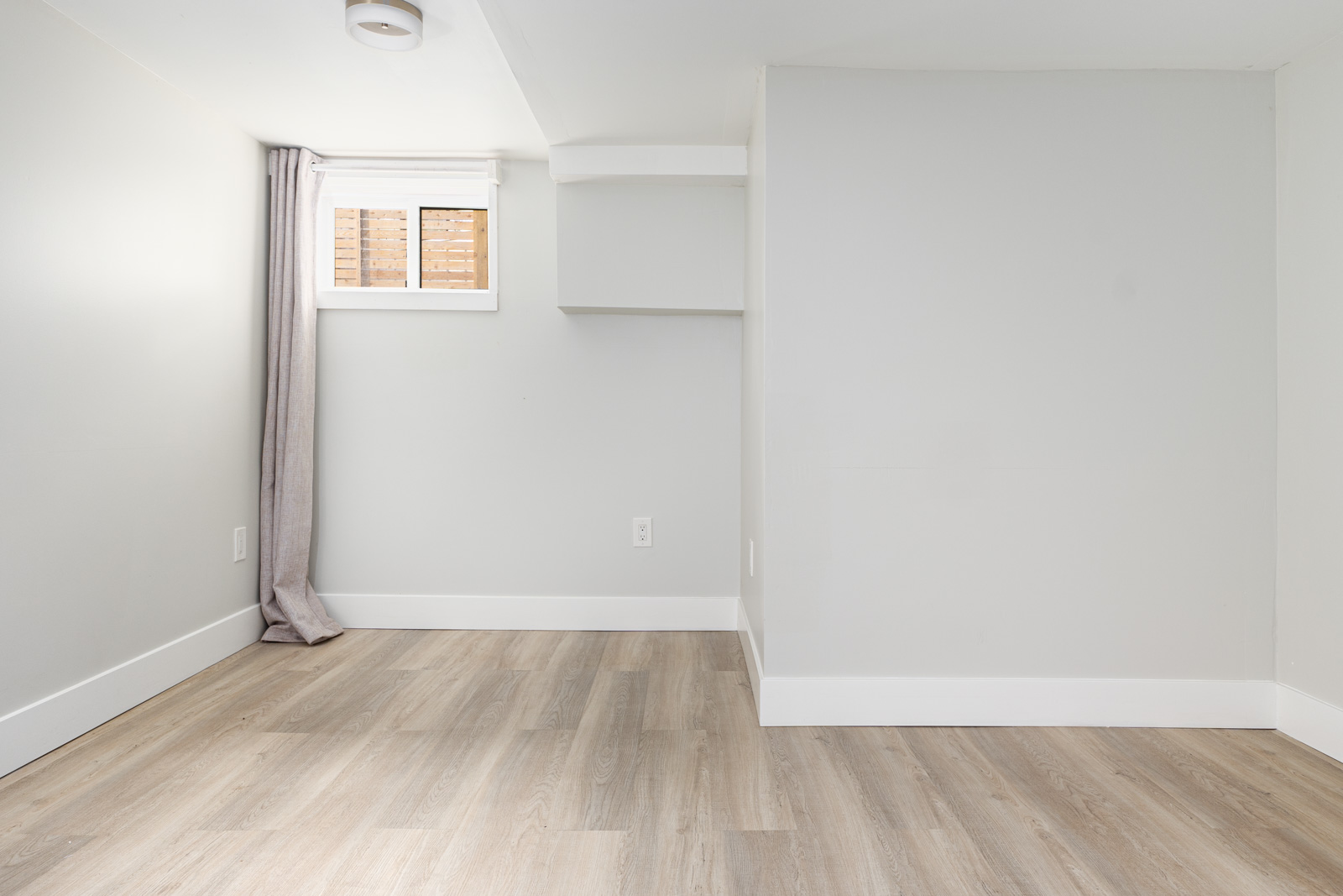 A small, empty room with light gray walls, a wood-patterned floor, a small window with gray curtains, and white baseboards.