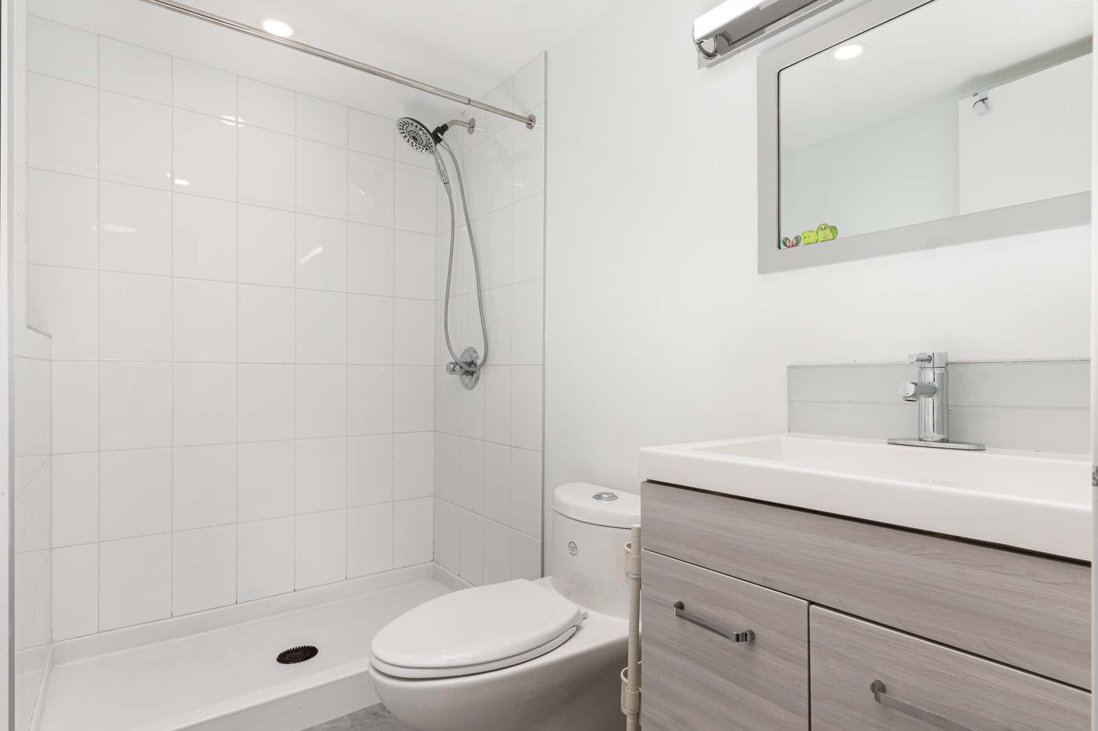 Modern bathroom with a walk-in shower, wall-mounted showerhead, toilet, and light wood vanity with a sink and mirror above. White tile and walls create a clean look.