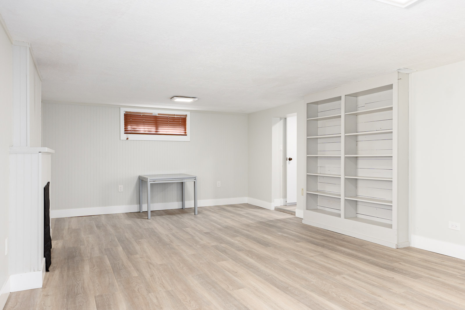 A clean, empty basement room with light wood flooring, built-in shelves, a small table, and a window with red blinds.