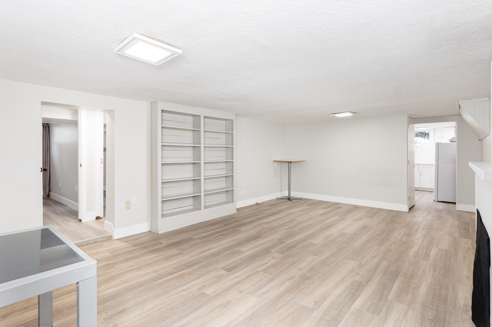 A spacious, unfurnished room with light wood flooring, built-in shelves, a small table, and visible entryways to other rooms including a kitchen.