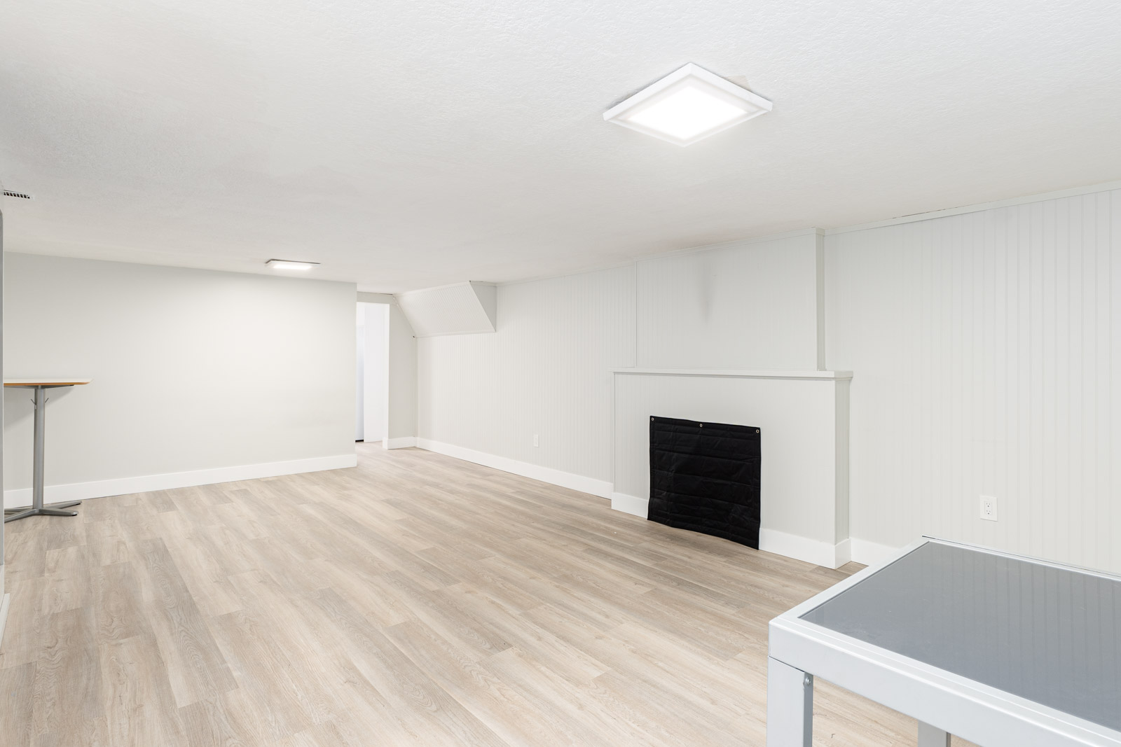 A spacious, unfurnished basement room with light wood flooring, white walls, a black fireplace, recessed lighting, and a small table in the corner.