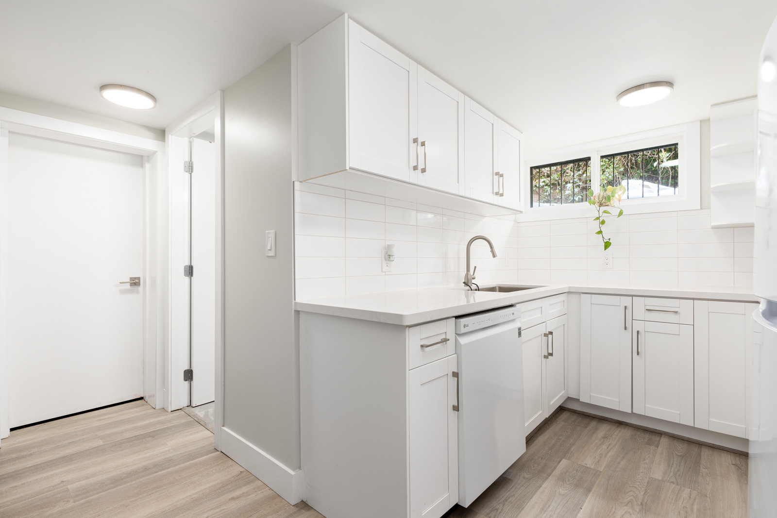 Modern kitchen with white cabinets, white tile backsplash, stainless steel sink, dishwasher, and light wood flooring. Small window above sink lets in natural light.
