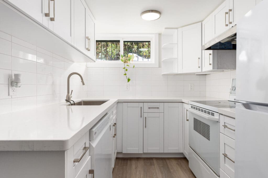 Modern white kitchen with upper and lower cabinets, stainless steel sink, electric stove, refrigerator, white tile backsplash, and a small plant by the window.