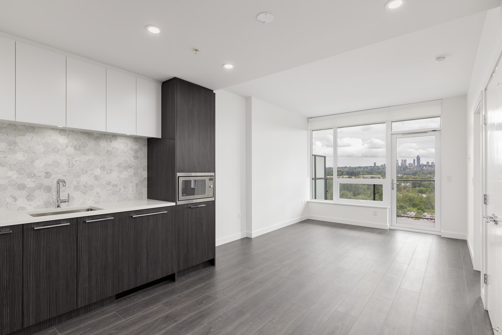 Modern kitchen and living area with dark wood cabinets, white countertops, and large windows offering a city view under a cloudy sky.
