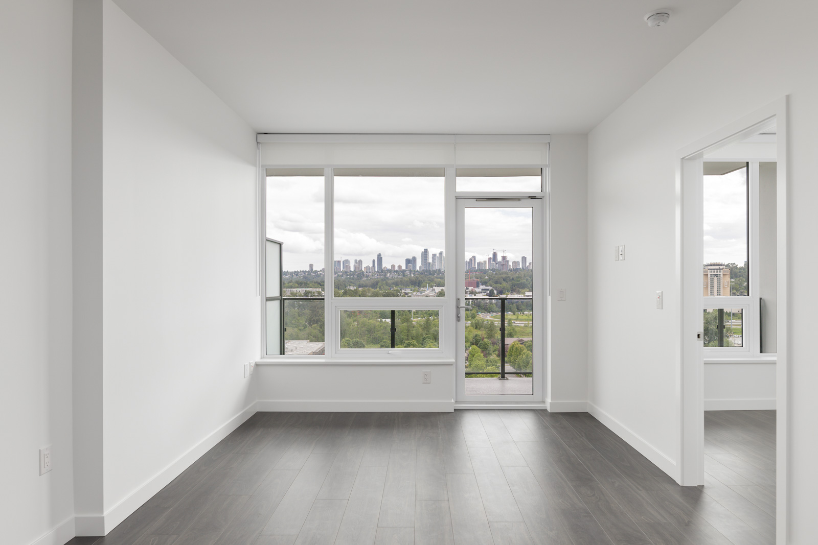 Unfurnished apartment room with large windows, balcony door, and a city skyline view; grey wood floors and white walls.