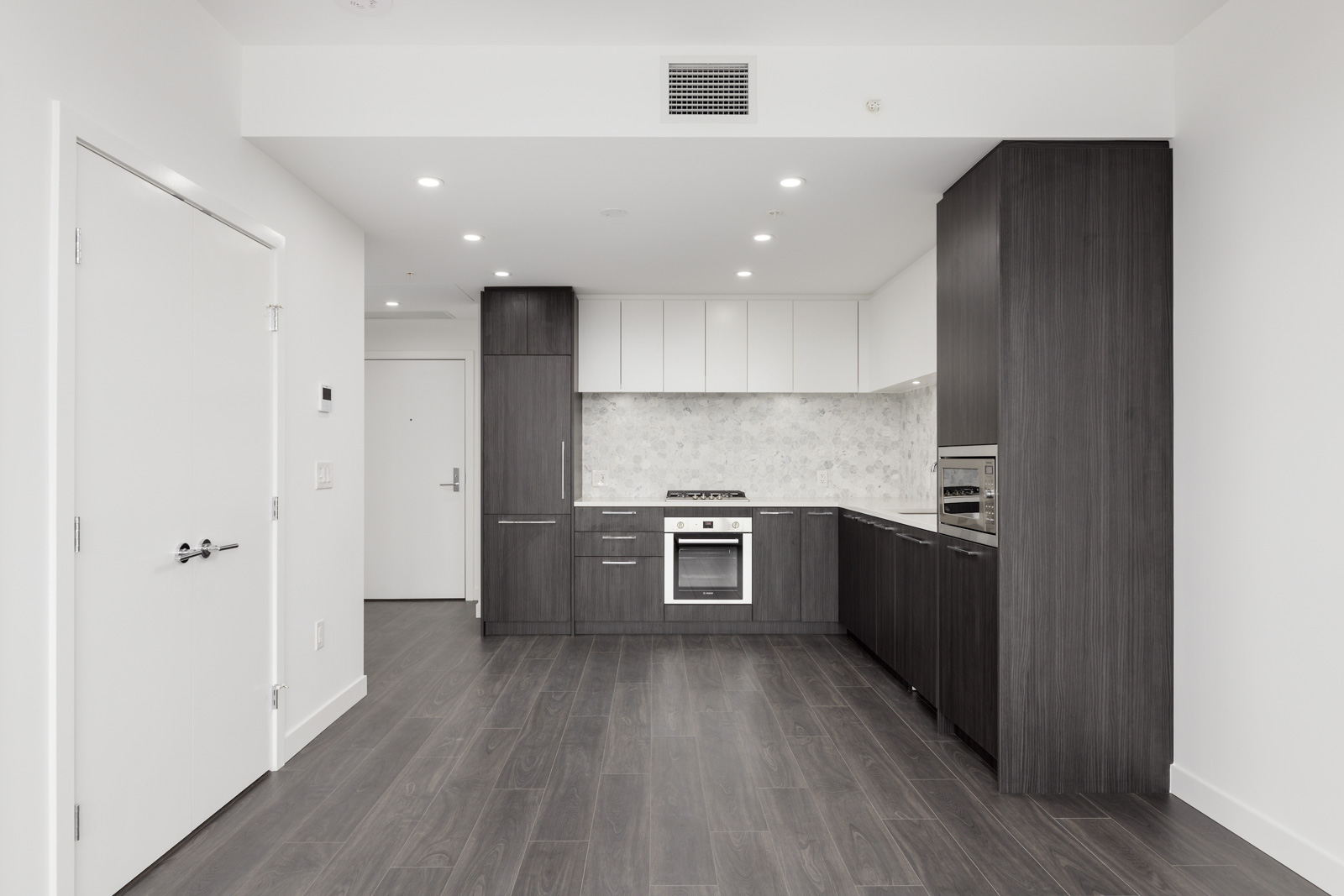 Modern kitchen with dark wood cabinets, white upper cabinets, built-in oven, gas stove, and grey wood flooring in a bright, minimalist apartment.