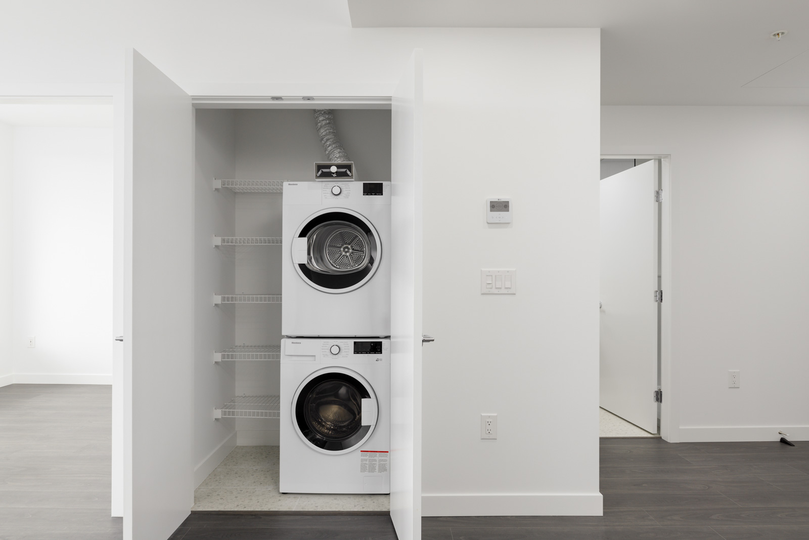 A small laundry closet with stacked front-loading washer and dryer, open double doors, and shelving beside the units in a modern white room.