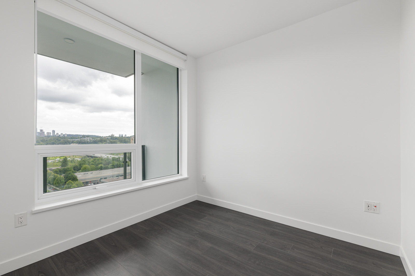 Empty white room with dark wood flooring, large floor-to-ceiling window, and a view of a city skyline and greenery outside.