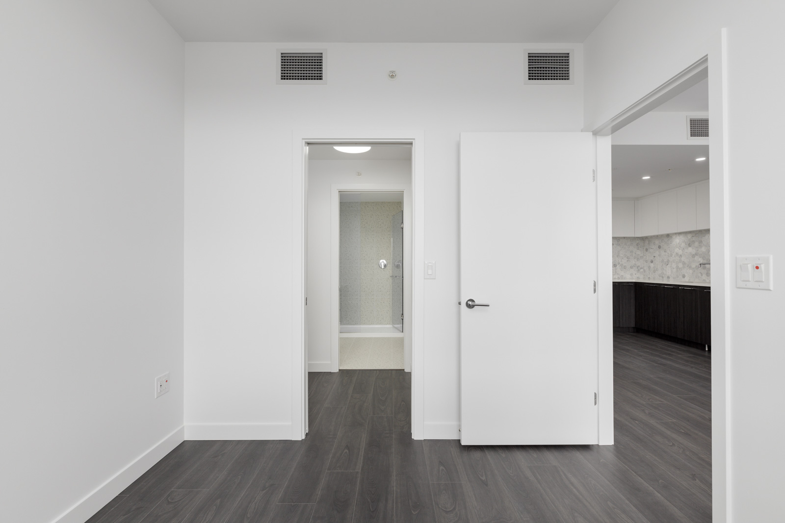 A white door opens to a hallway leading to a bathroom, with another doorway to a kitchen area on the right. The room has dark wood floors and white walls.