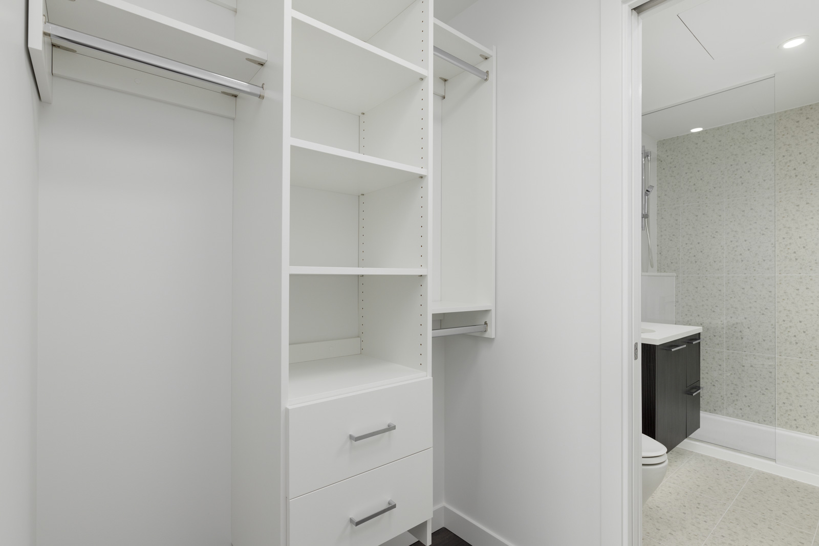 A white walk-in closet with shelves, drawers, and hanging rods, adjacent to a modern bathroom with a vanity and toilet.