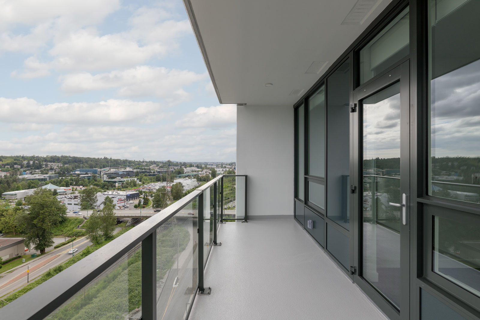 A modern apartment balcony with glass railing overlooks a suburban area with roads, trees, buildings, and a cloudy sky in the distance.