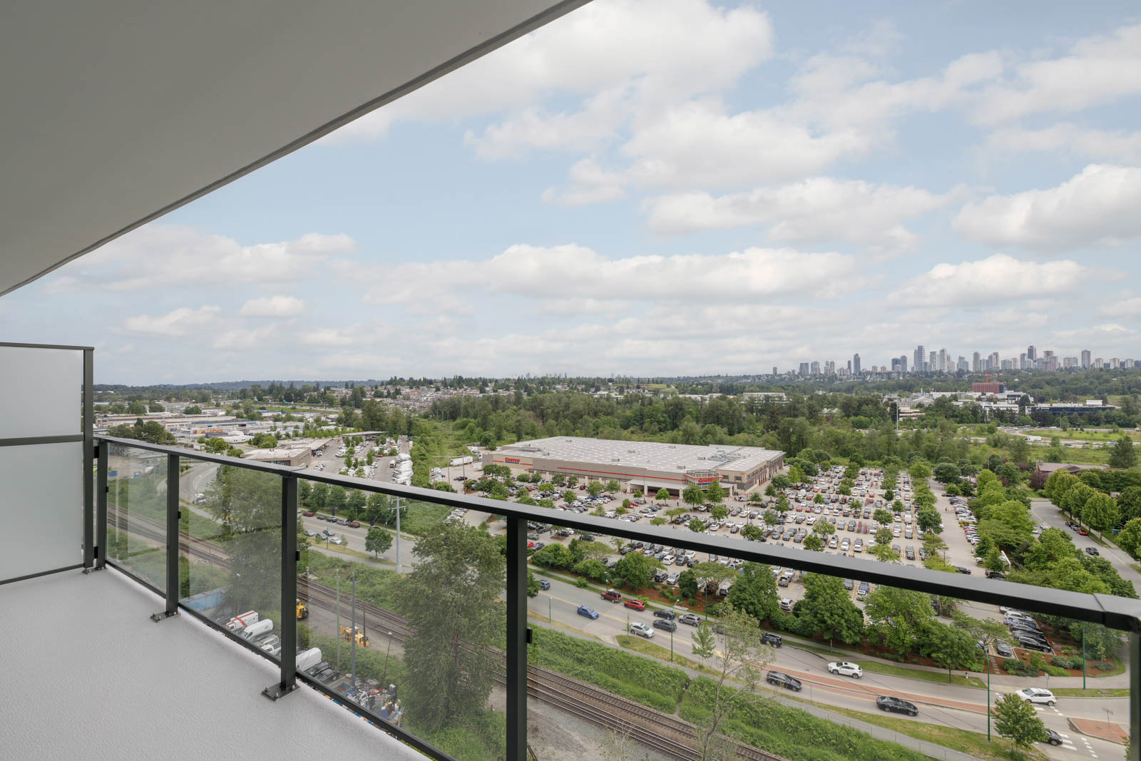 View from a high-rise balcony overlooking a large parking lot, commercial buildings, trees, and a distant city skyline under a partly cloudy sky.