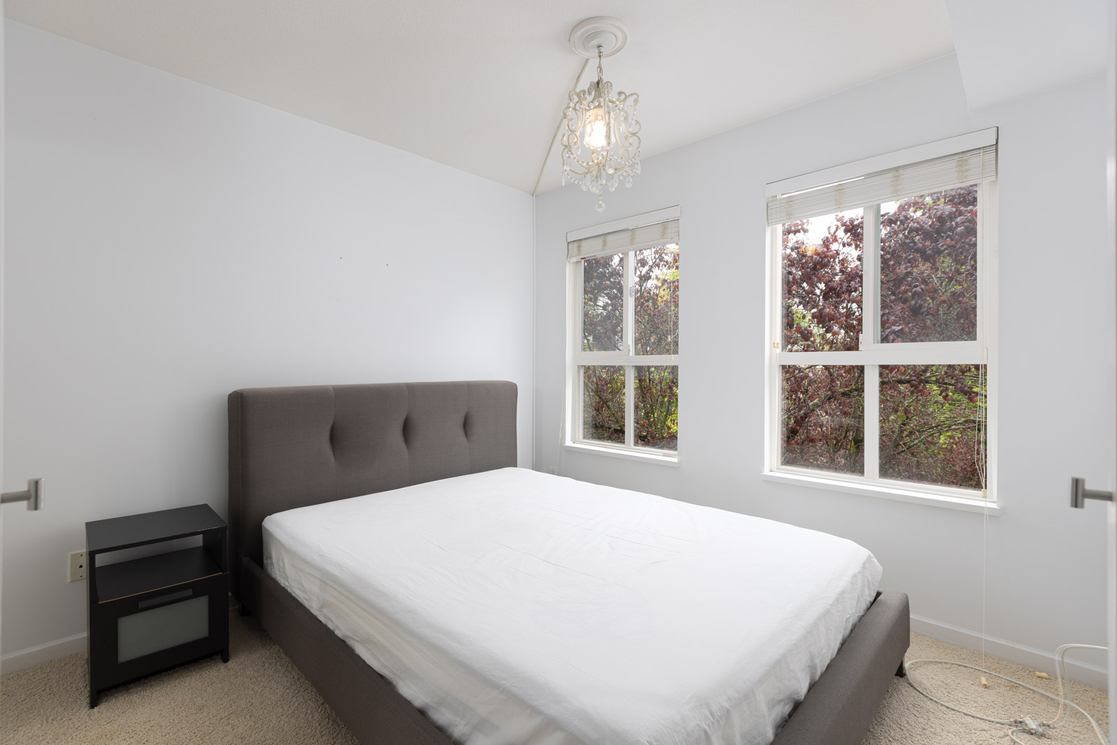 A minimally furnished bedroom with a gray upholstered bed frame, white bedding, a black nightstand, two windows, and a ceiling light fixture.