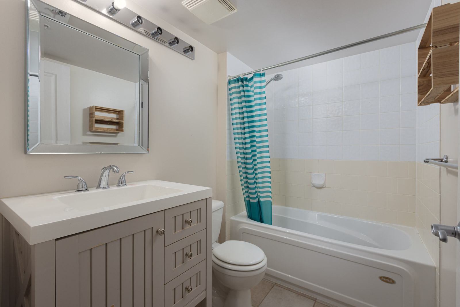 Bathroom with a white vanity, mirror, toilet, bathtub with a blue-striped shower curtain, and wall shelves. White tile walls and neutral floor tiles.