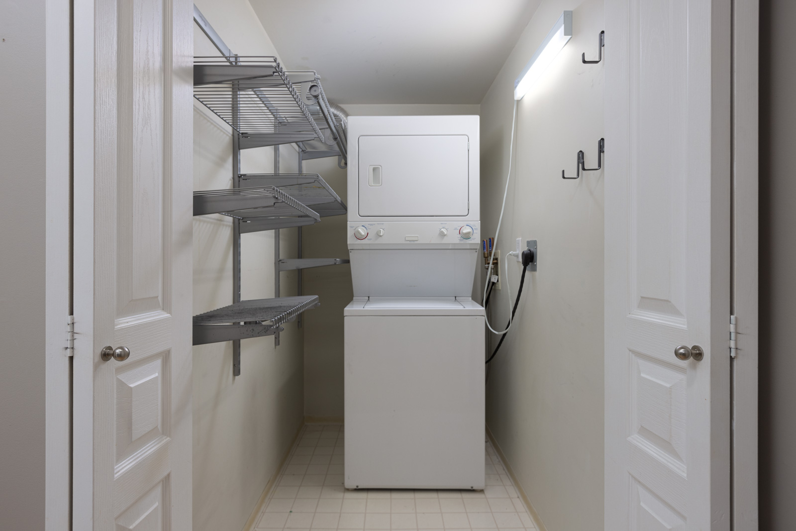 A small laundry closet with a stacked washer and dryer, wire shelving on the left wall, and hooks on the right wall, illuminated by a fluorescent light.