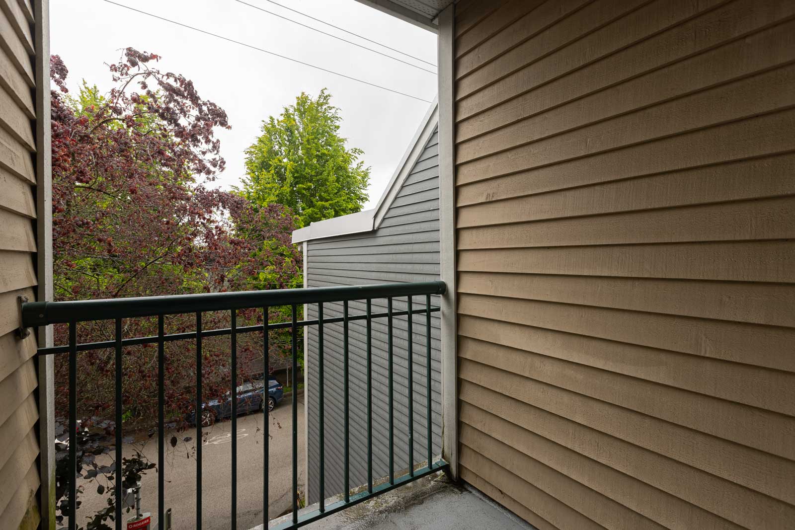 View from a small balcony with beige siding, metal railing, and a partial view of trees, rooftops, and parked cars on a street below.