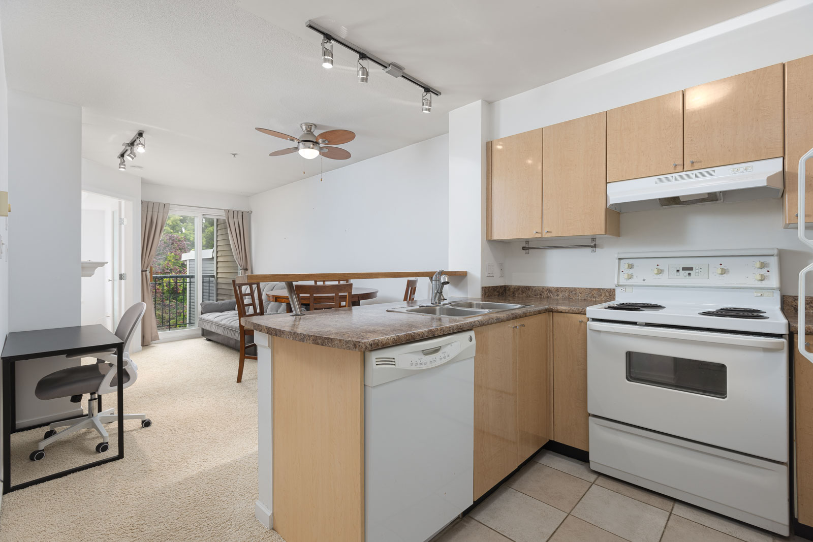 Modern apartment kitchen with light wood cabinets, white appliances, and adjacent dining and living area; sliding glass door leads to a balcony with greenery outside.