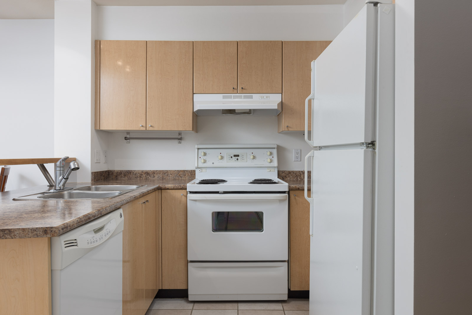 A small kitchen with light wood cabinets, a white stove and range hood, a white refrigerator, a dishwasher, and a double sink with a brown countertop.