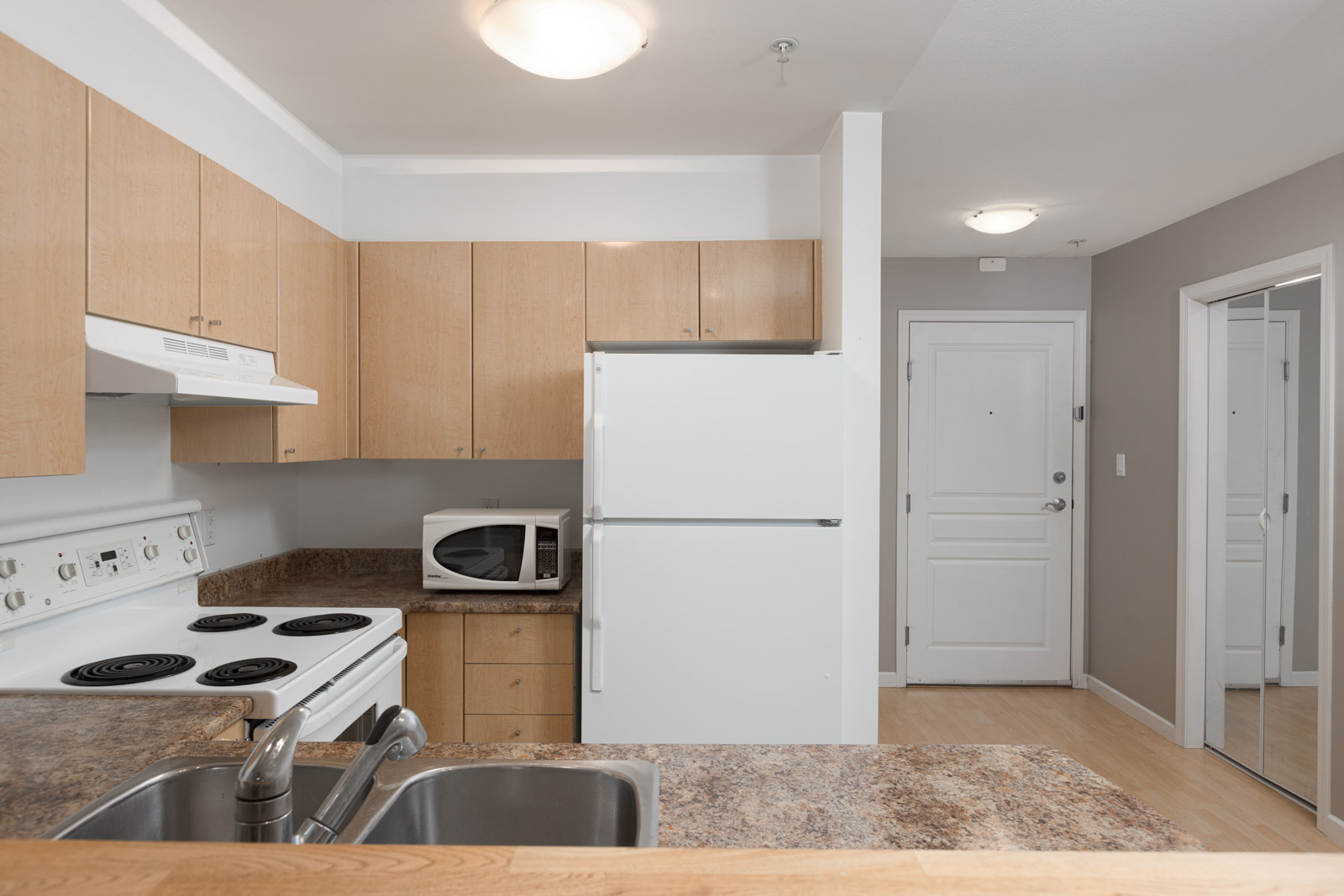 A modern kitchen with light wood cabinets, white appliances including a stove, microwave, and refrigerator, and a double sink in the foreground.