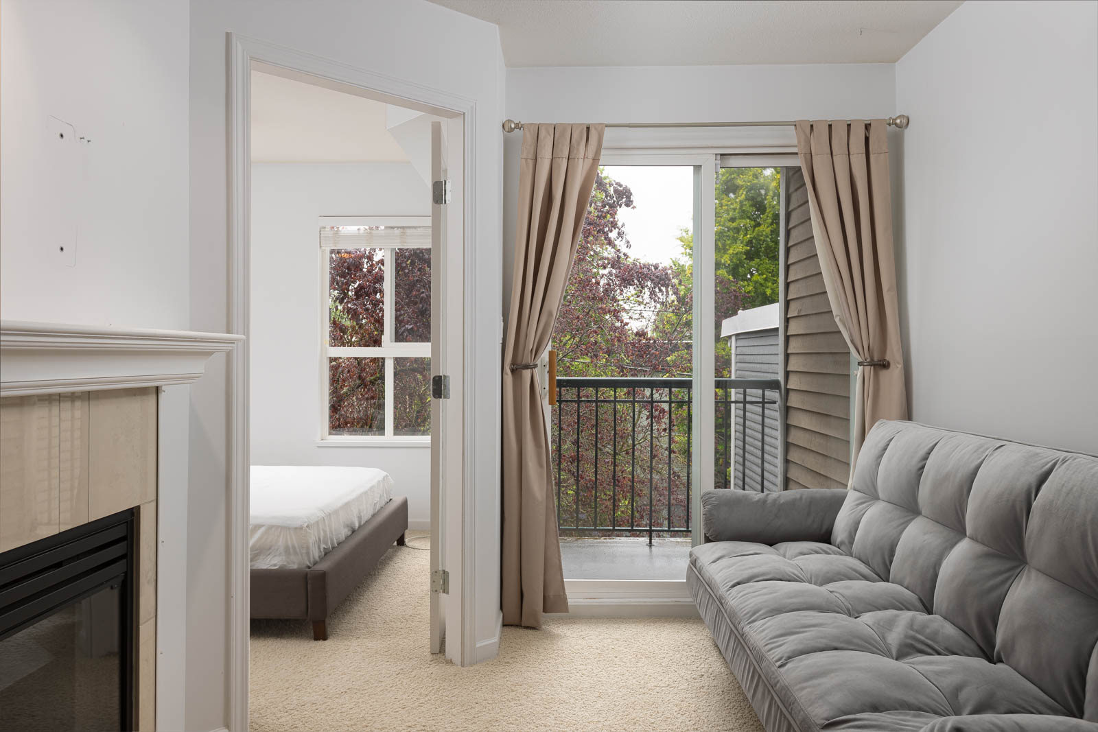 A living room with a gray sofa, fireplace, and glass doors opening to a small balcony; a bedroom with a bed is visible through an open door.