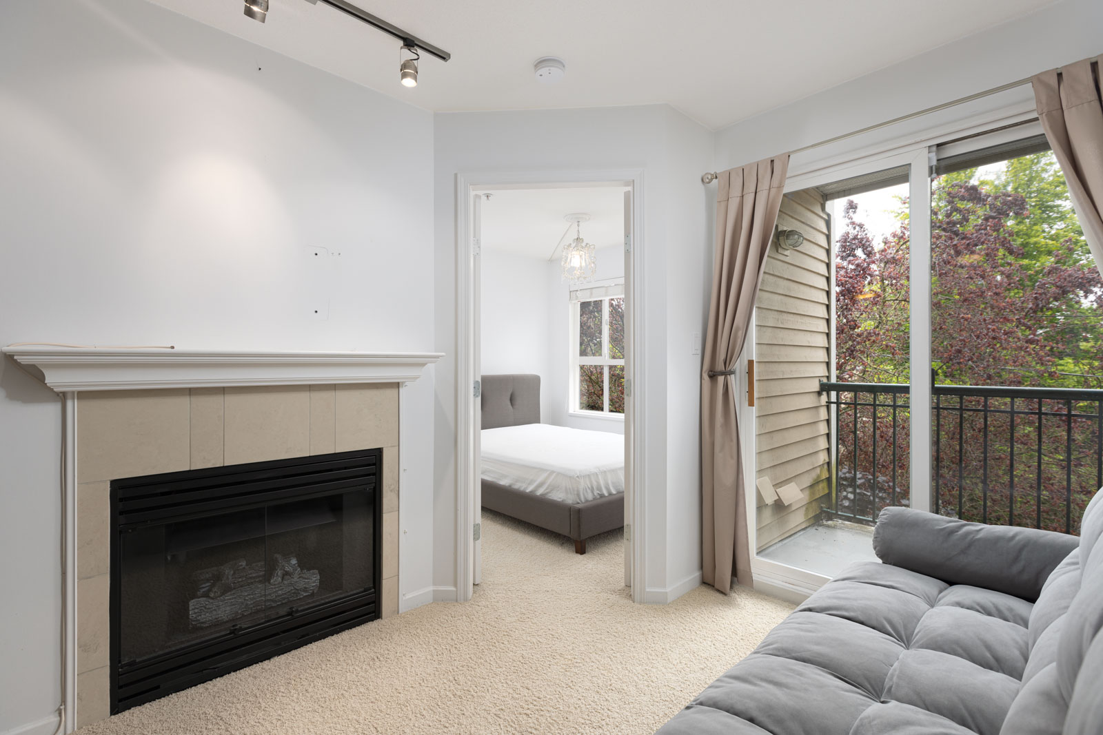 Living room with a fireplace, gray sofa, and sliding glass doors to a balcony. Bedroom with a bed and window visible through an open doorway.