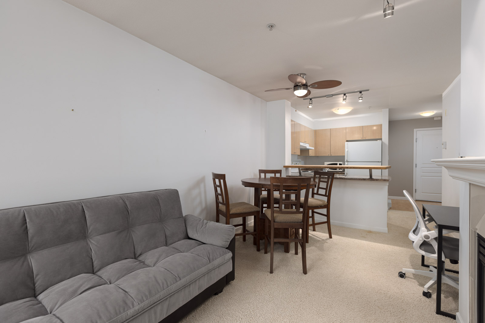 A living area with a gray futon, a round dining table with four chairs, an open kitchen, and a white desk with a chair near a fireplace.