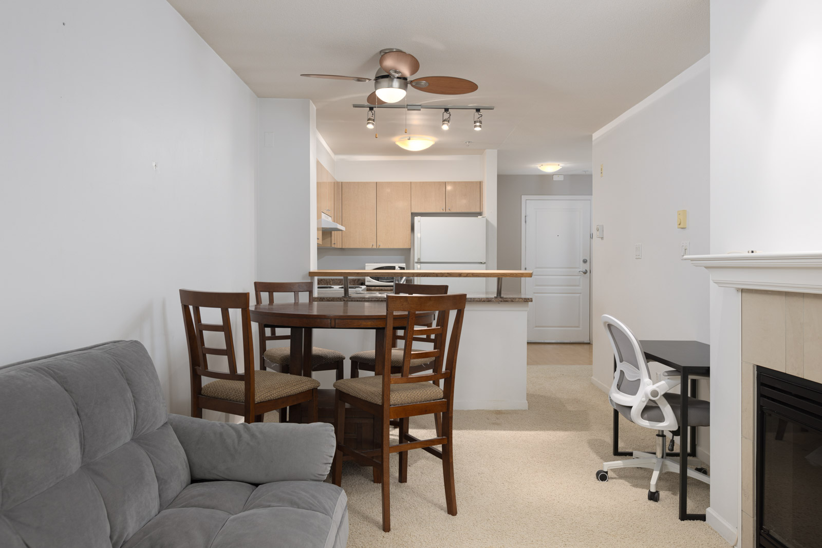 A small apartment dining area with a round wooden table and four chairs, adjacent to a kitchen, a gray couch, a white desk chair, and a ceiling fan with lights.