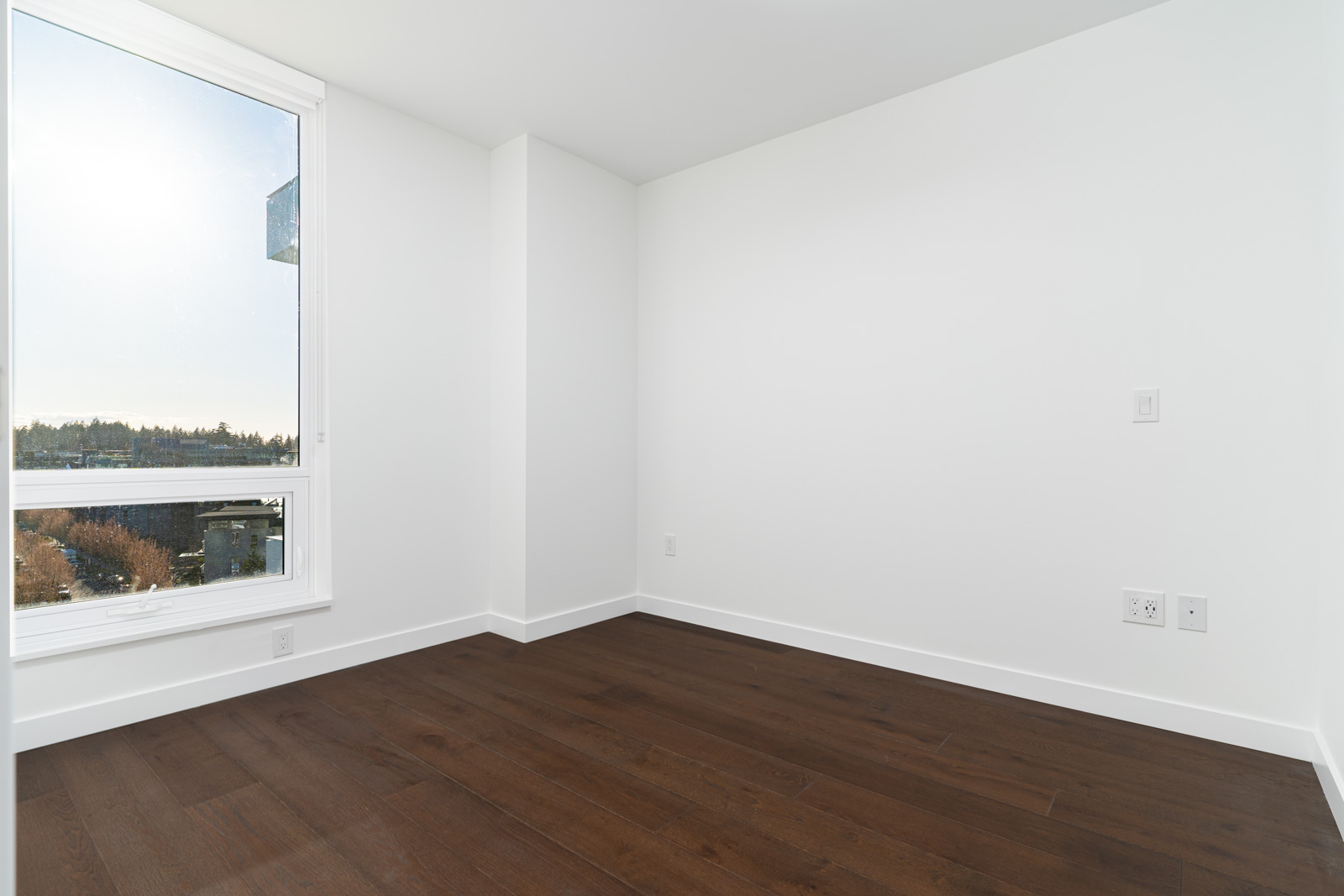 Empty room with white walls, a large window on the left letting in daylight, and dark hardwood flooring. Electrical outlets are visible on the right wall.