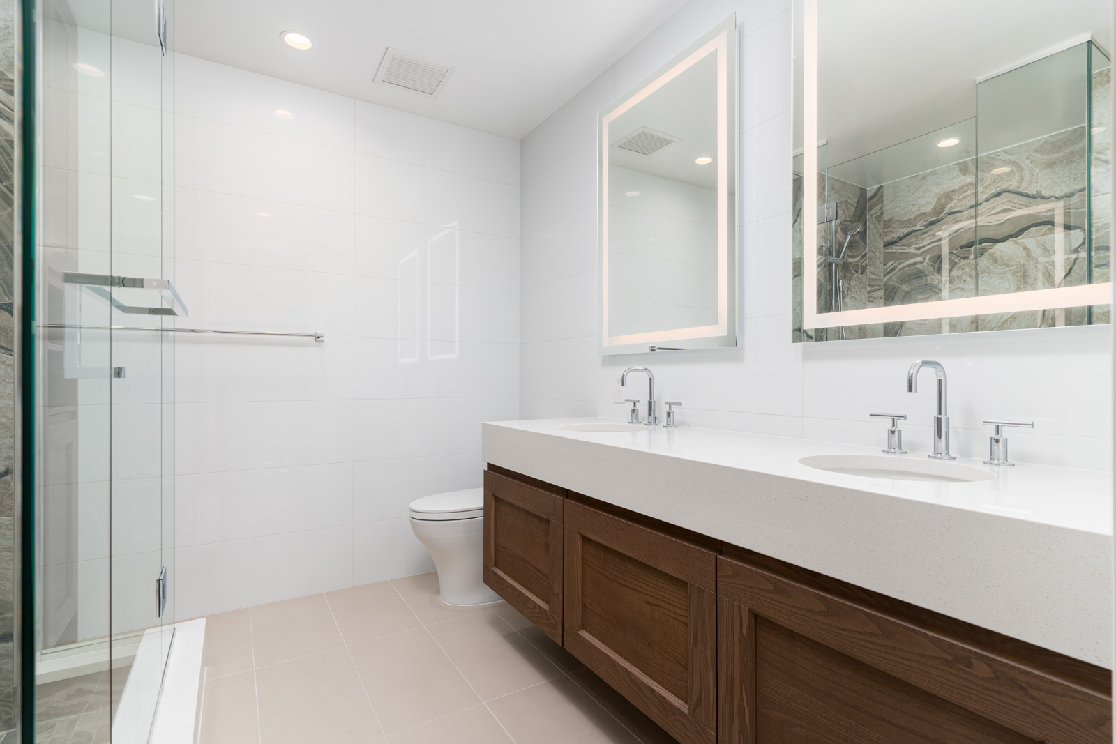 Modern bathroom with dual sinks, large illuminated mirror, wooden cabinetry, glass shower enclosure, and white tile walls and flooring.