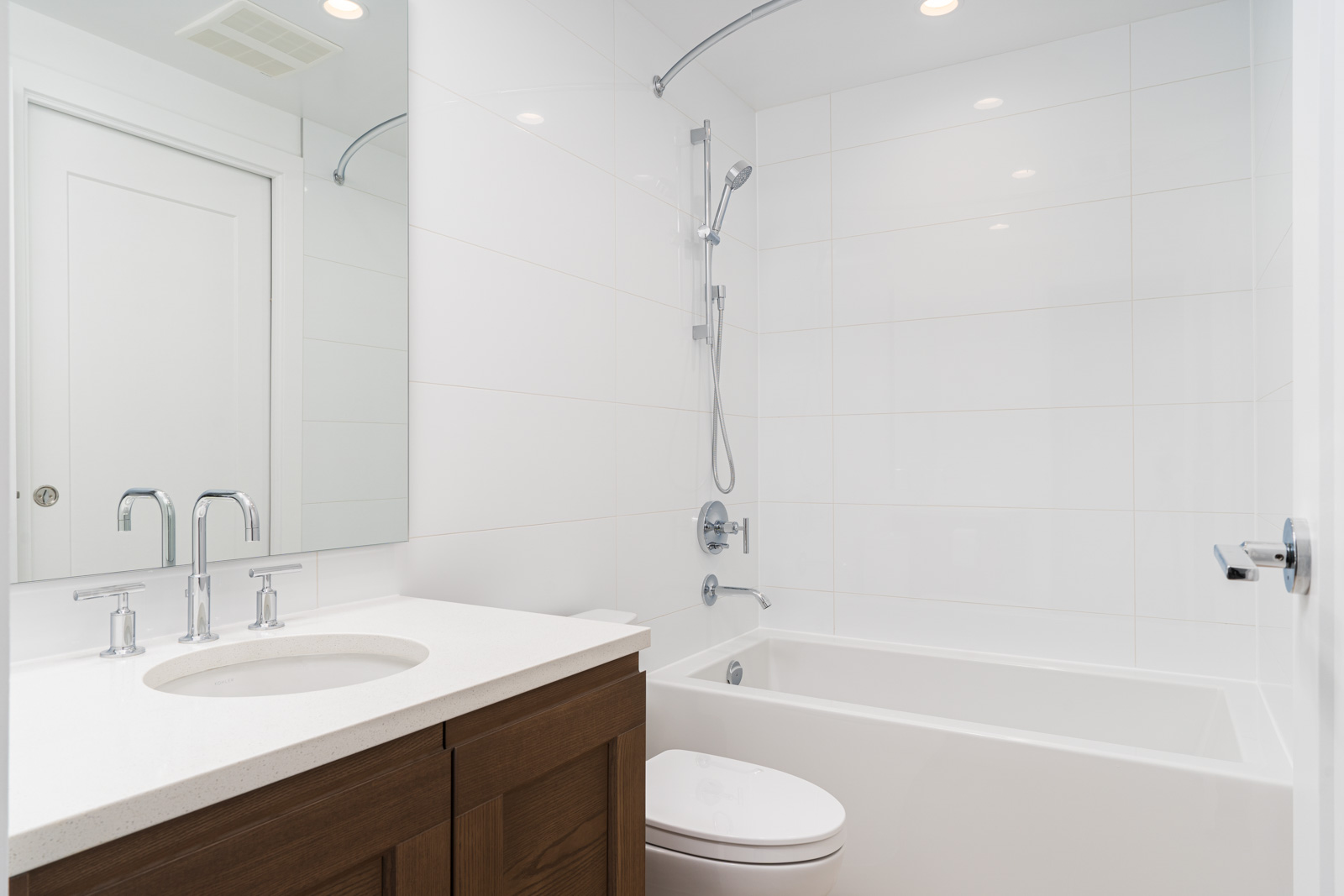 Modern bathroom with a white bathtub, wall-mounted showerhead, toilet, white countertop with sink, and wooden vanity. White tiled walls and ceiling lights provide a clean, bright appearance.