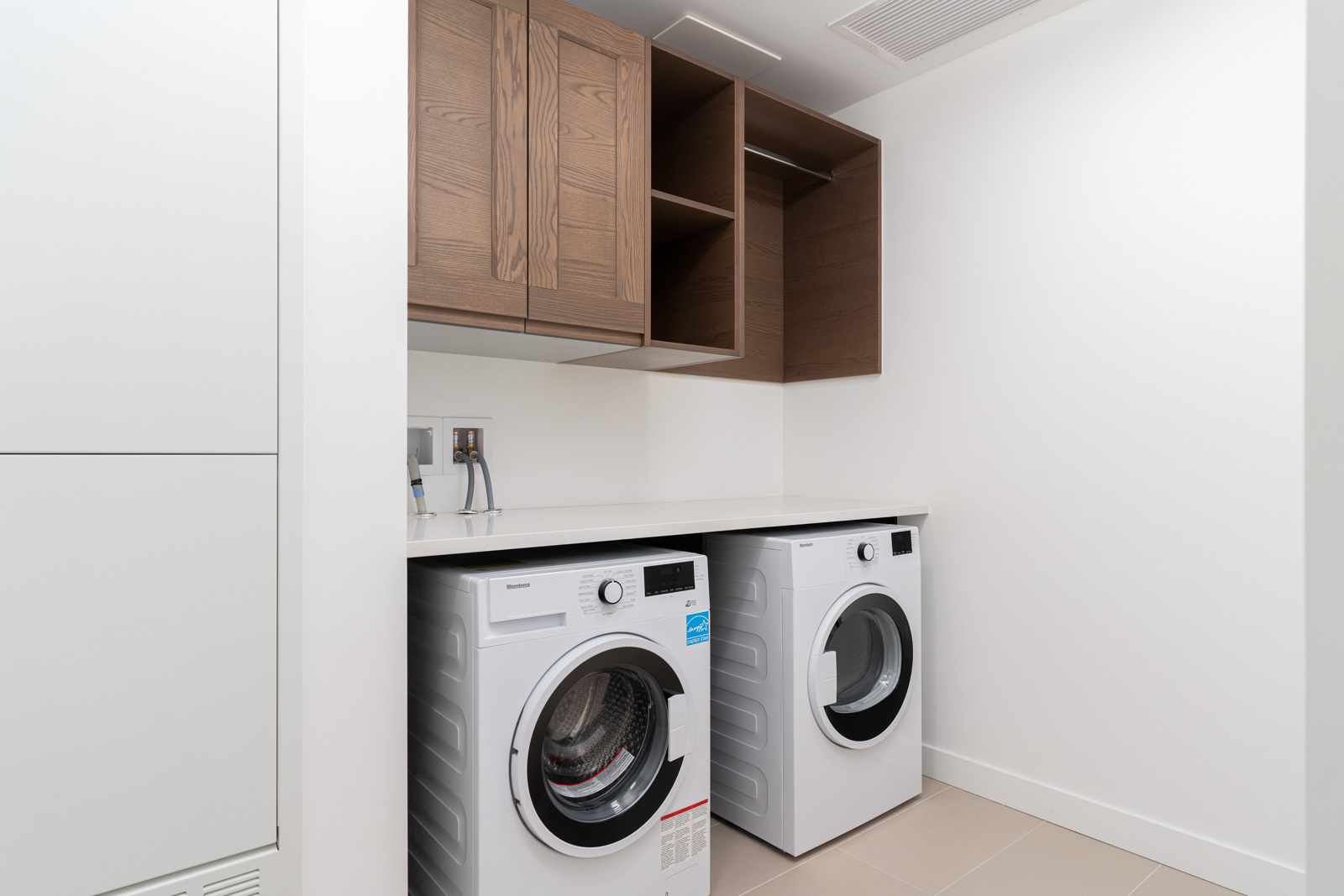 A modern laundry room with a front-loading washing machine and dryer, wooden overhead cabinets, and a white countertop.