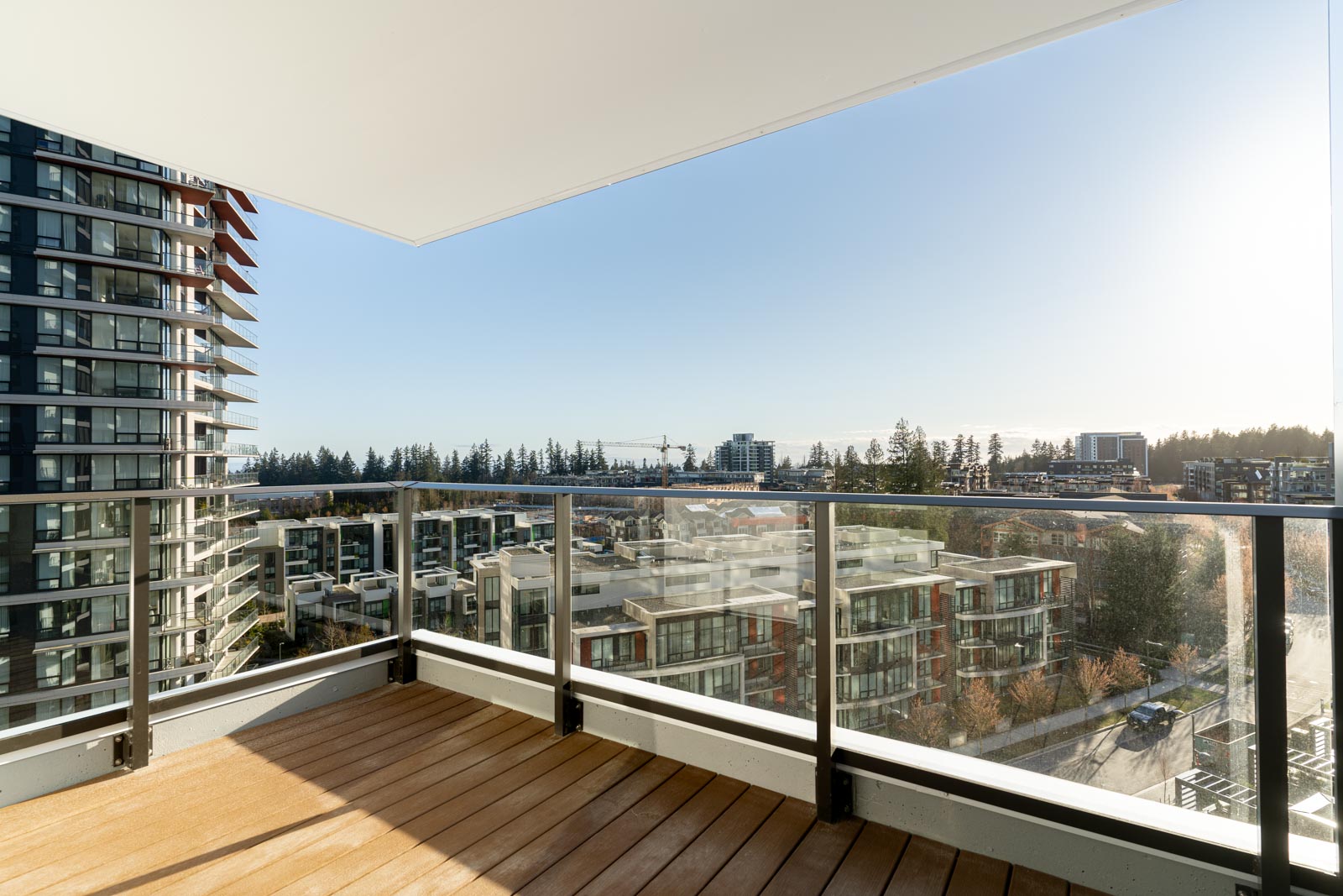 Glass-railed balcony with wooden floor overlooking modern apartment buildings and a distant cityscape under a clear blue sky.