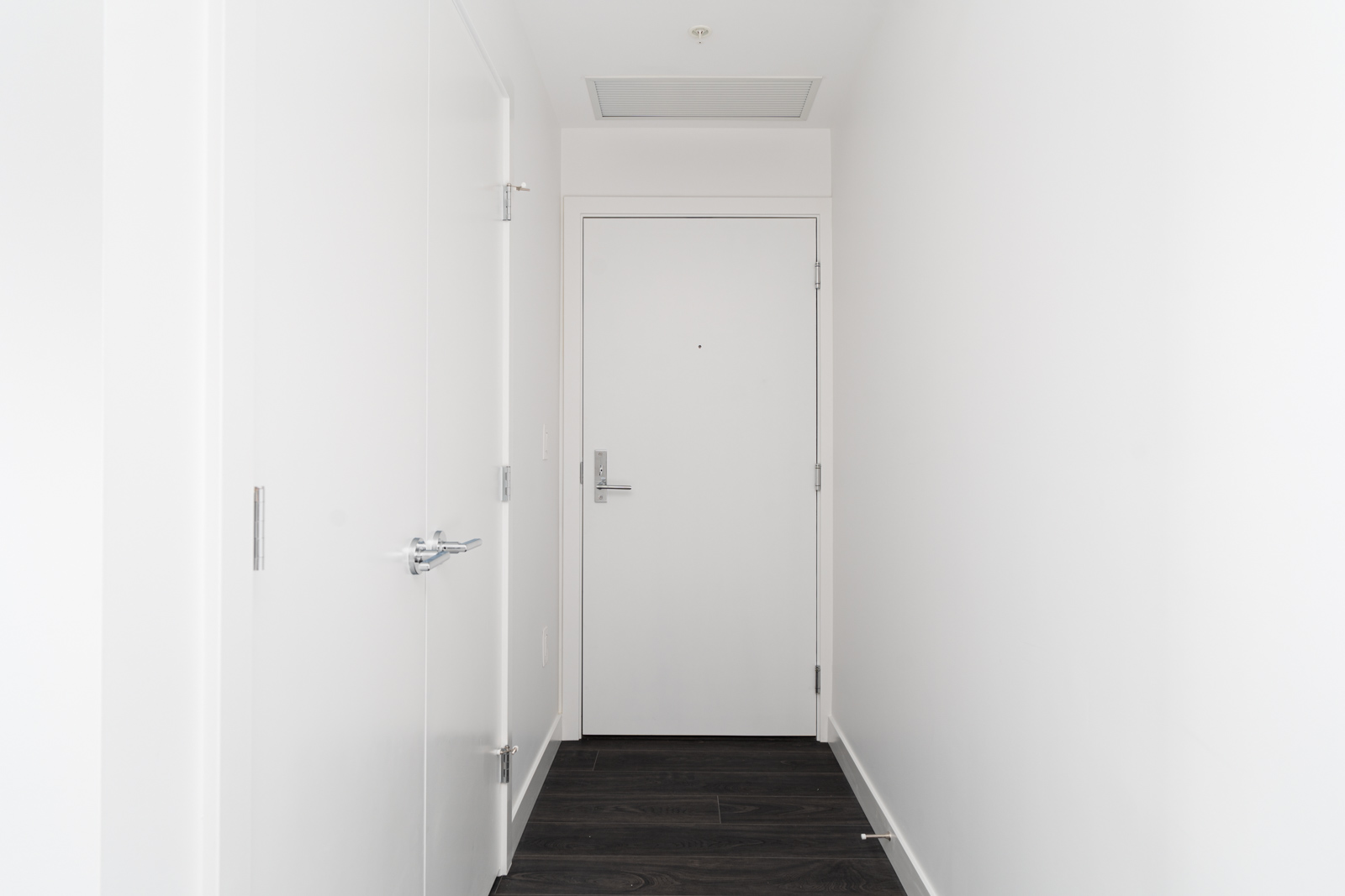 A narrow hallway with white walls and a white door at the end, featuring dark wood flooring and a closed side door on the left.
